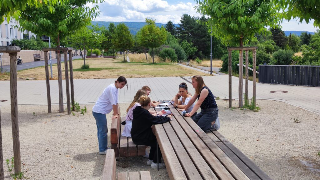 A group of people sit together at a table in the middle of a sunlight square. They are discussing the attributes of their surroundings and are filling out surveys.