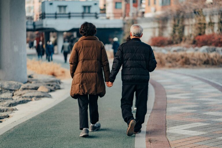 Older couple walking hand in hand on a path in an urban area. Both wear dark winter jackets and pants. Other people are walking in the background. The path curves beside rocks and dry plants, with buildings and a bridge visible ahead.