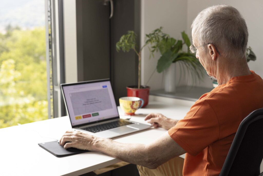 Older man sitting at a desk and using a laptop to participate to the Silver Ways Survey. A window with a view of green trees is open in the background.