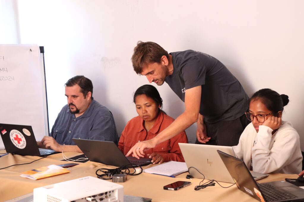 Four people sitting and standing around a table with laptops, notebooks, and electronic devices in a meeting or collaborative work setting.