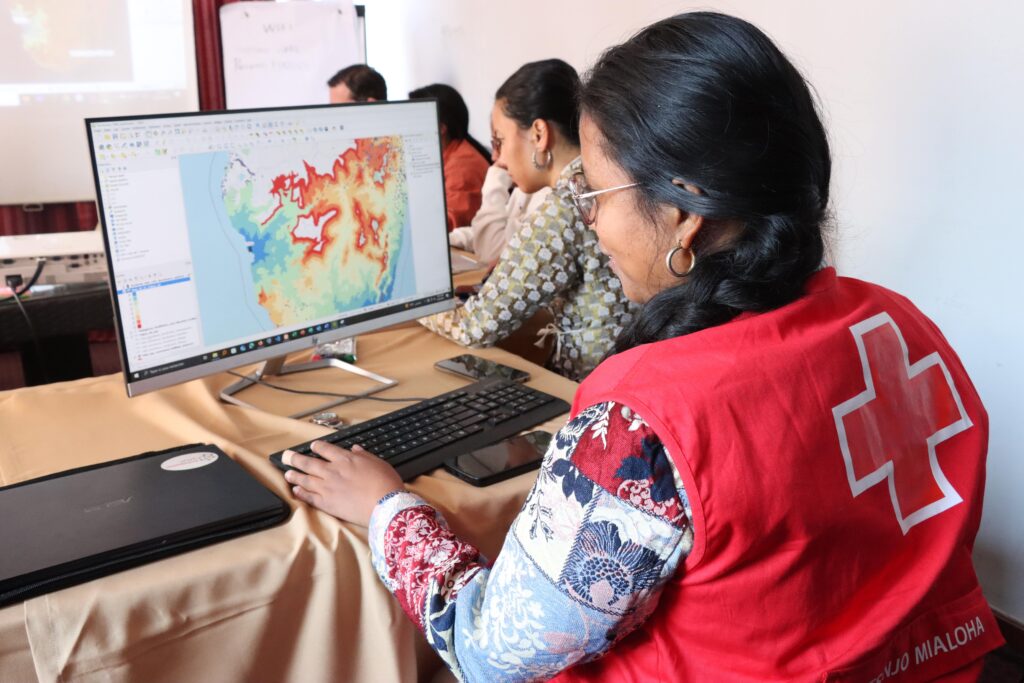 Person wearing a red vest with a white cross logo working on a computer displaying a colorful topographic map with elevation data.