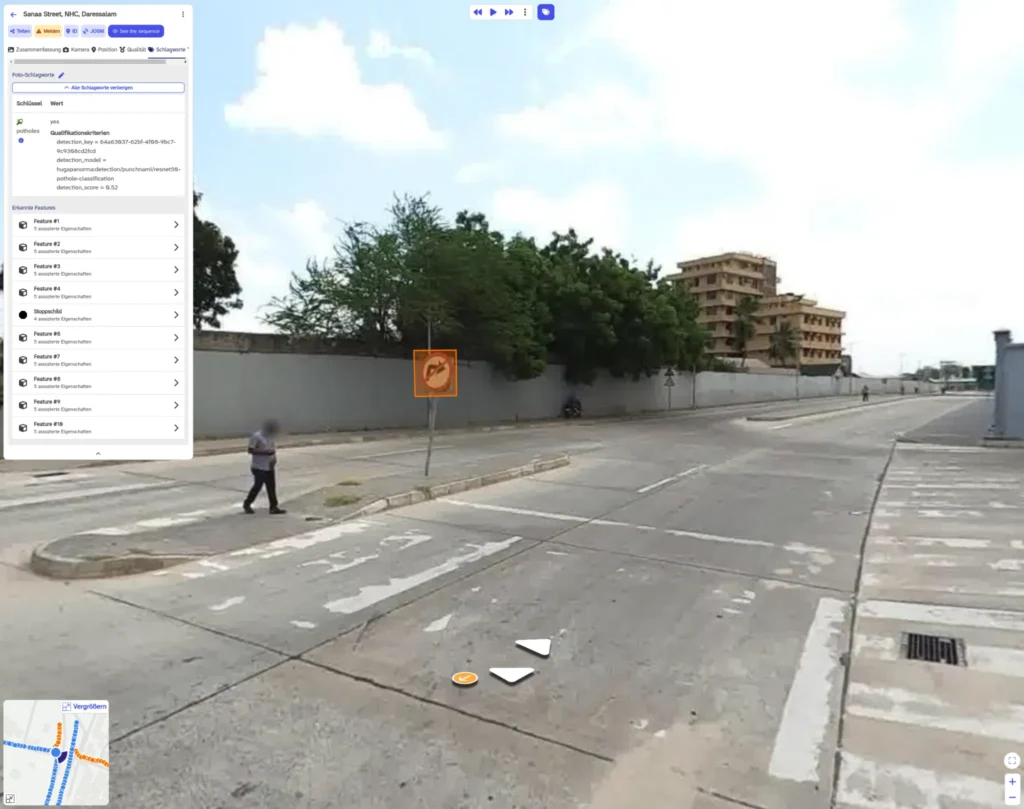 Man walking across a wide concrete street with faded crosswalk markings, trees and a multi-story building in the background.