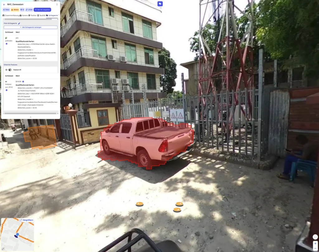 Red pickup truck parked on a sunlit street near a multi-story building and a metal tower with fencing.