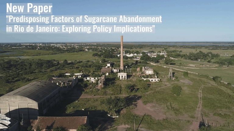 Aerial view of an abandoned sugarcane processing facility with a tall chimney and surrounding green fields under a clear sky.