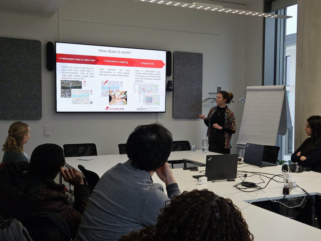 A group of people are sitting in a conference room listening to a presentation about the Sketch Map Tool.