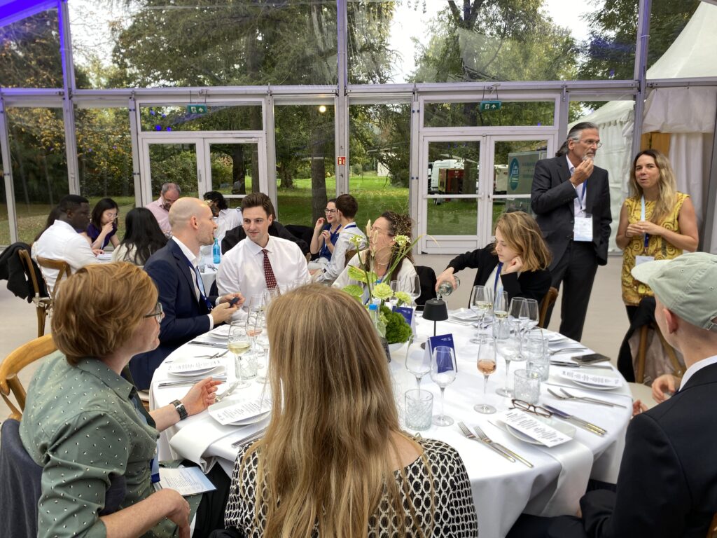 Group of people seated and standing around a round table set with glasses, plates, and cutlery in a glass-walled room with greenery outside.