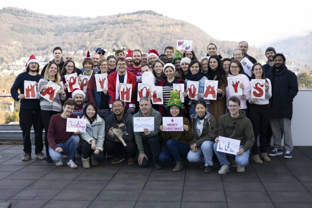 Group of people outdoors holding signs that spell 'HAPPY HOLIDAYS' and other Christmas messages, some wearing Santa hats and reindeer antlers.