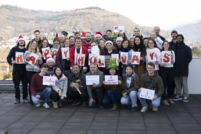 Group of people outdoors holding signs that spell 'HAPPY HOLIDAYS' and other Christmas messages, some wearing Santa hats and reindeer antlers.