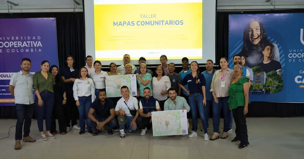 Group of people standing and kneeling in front of a yellow presentation screen with the text 'TALLER MAPAS COMUNITARIOS' and holding community maps