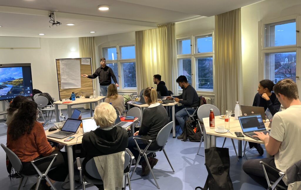 Mehrere Personen sitzen an Tischen mit Laptops in einem hellen Raum, während eine Person vorne an einem Whiteboard steht und auf einen Bildschirm zeigt.