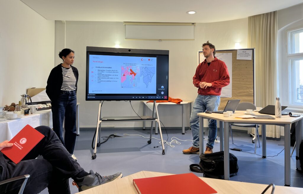 Zwei Personen stehen vor einem großen Bildschirm mit einer Präsentation, im Vordergrund sind Tische mit roten Ordnern und Laptops zu sehen.