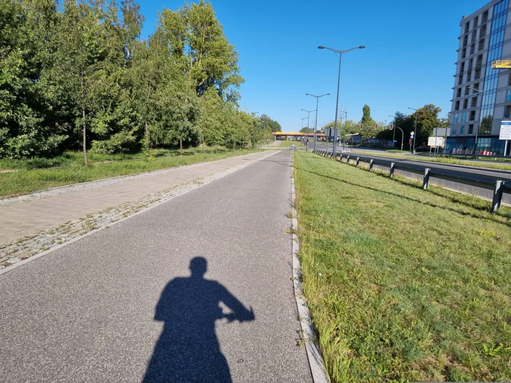 Bike path with shadow of a person on handlebars, trees on the left, grassy strip and road with guardrail on the right