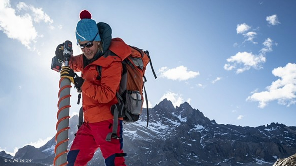 Mountaineer wearing red jacket and blue hat carrying equipment on back in front of mountain peaks under clear sky