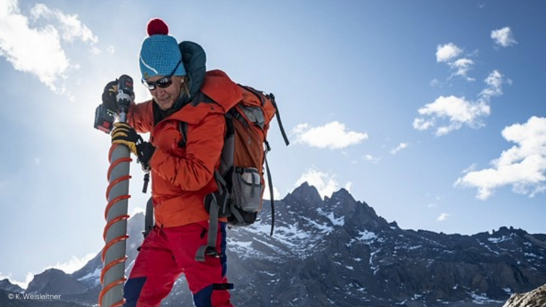 Mountaineer wearing red jacket and blue hat carrying equipment on back in front of mountain peaks under clear sky
