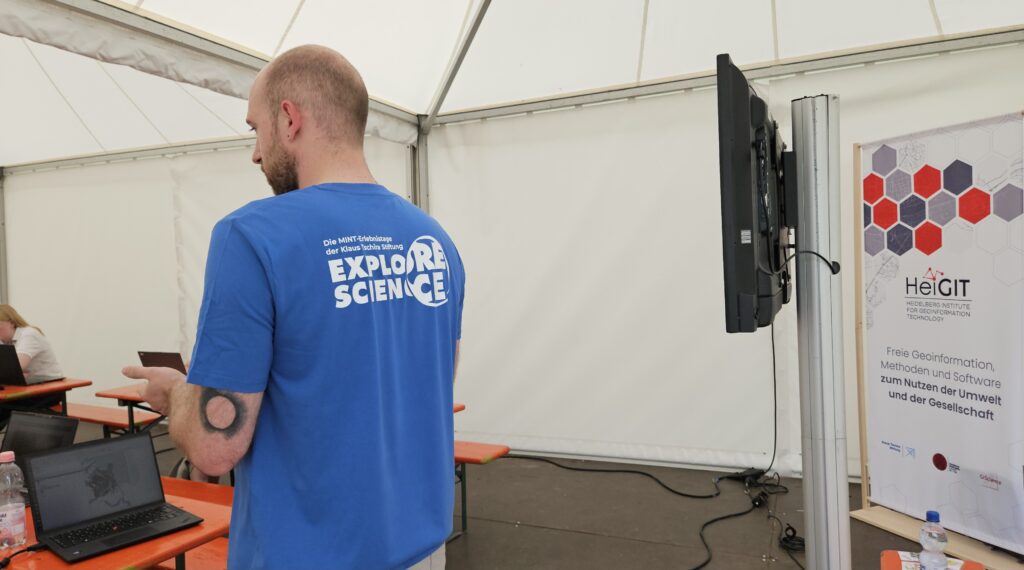 Man in blue 'Explore Science' T-shirt standing in front of a laptop and a monitor inside a tent with a poster about HeiGIT on the right