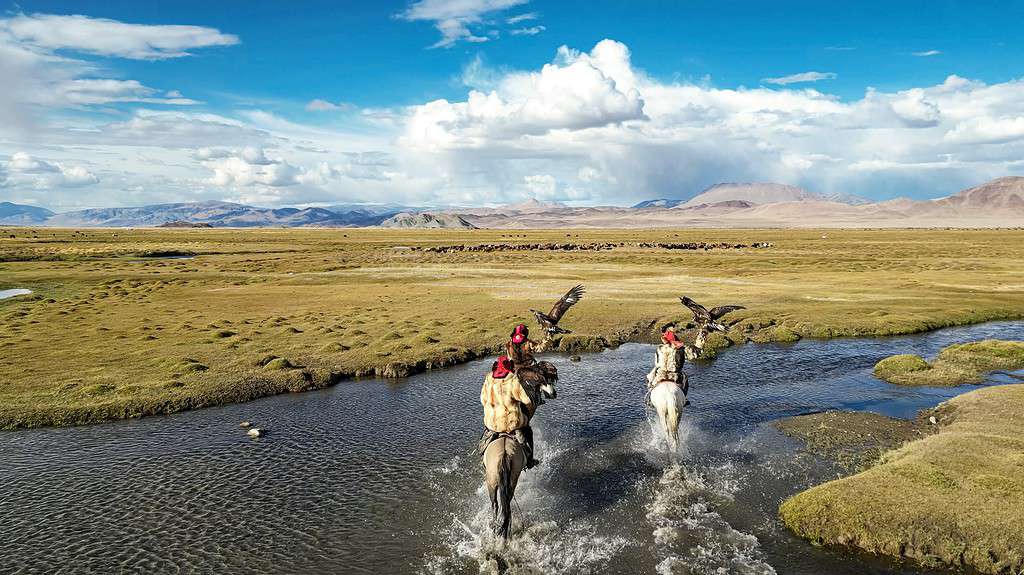 Two riders on horseback crossing a shallow river in a wide open plain under a partly cloudy sky