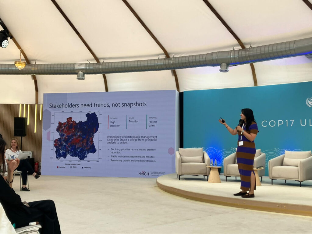 Woman in striped dress presenting on stage with a large screen showing a map and text about stakeholder needs